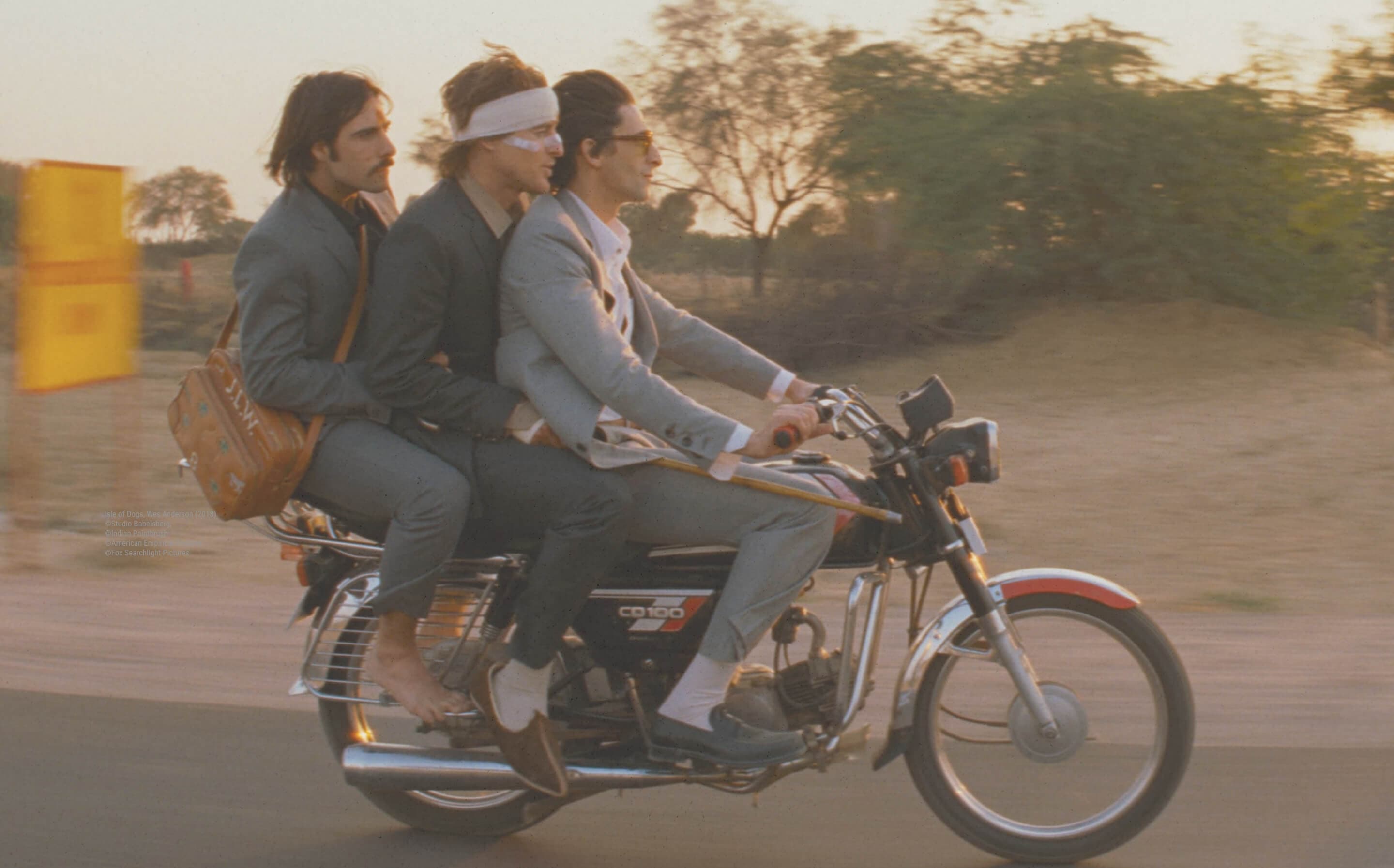 Three men in grey suits riding one motorcycle down a dusty road at sunset